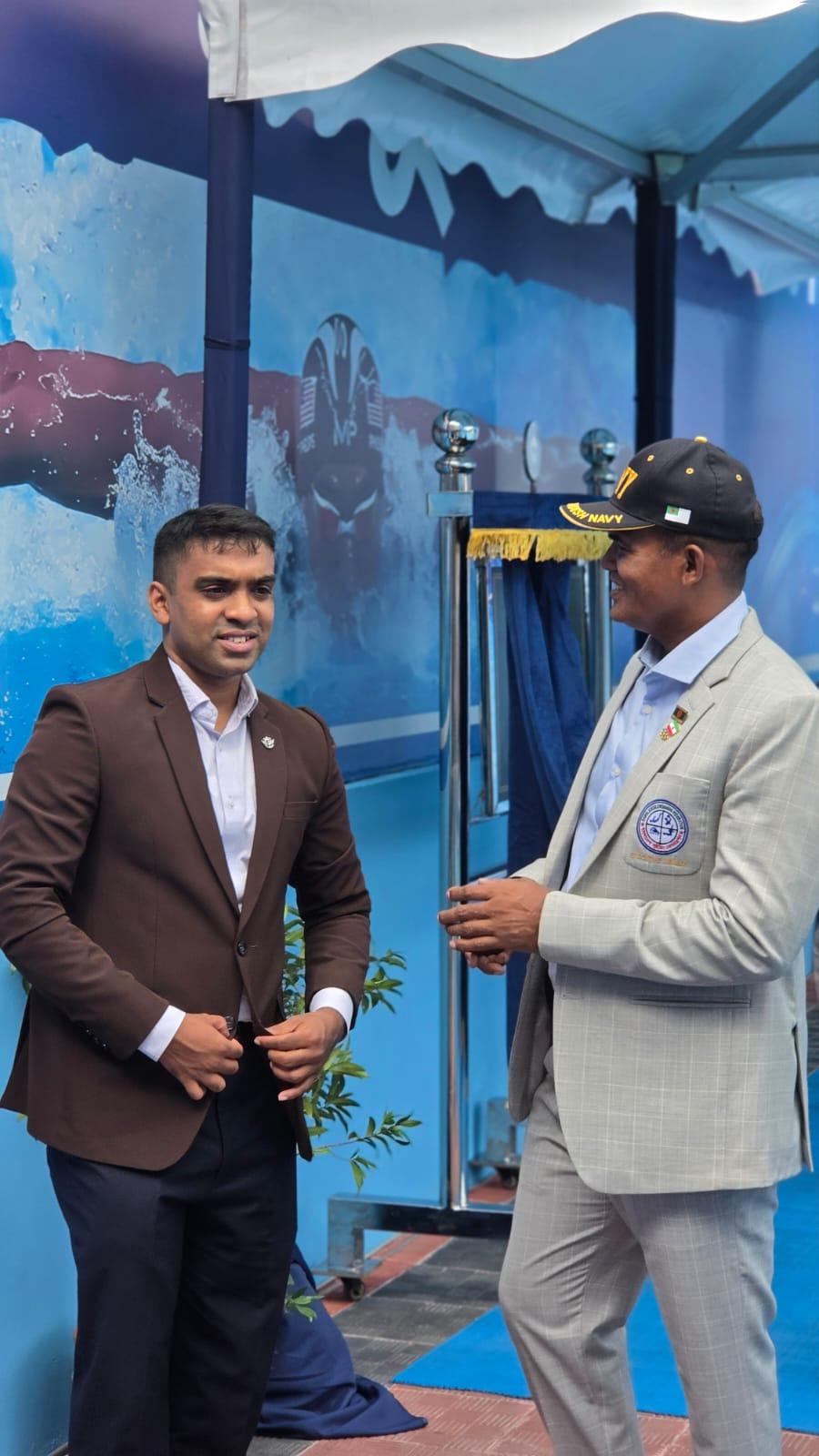 A Bangladesh Navy officer in a cap and blazer talks with another guest beside the ribbon-cutting stage, its ocean-mural backdrop behind them.