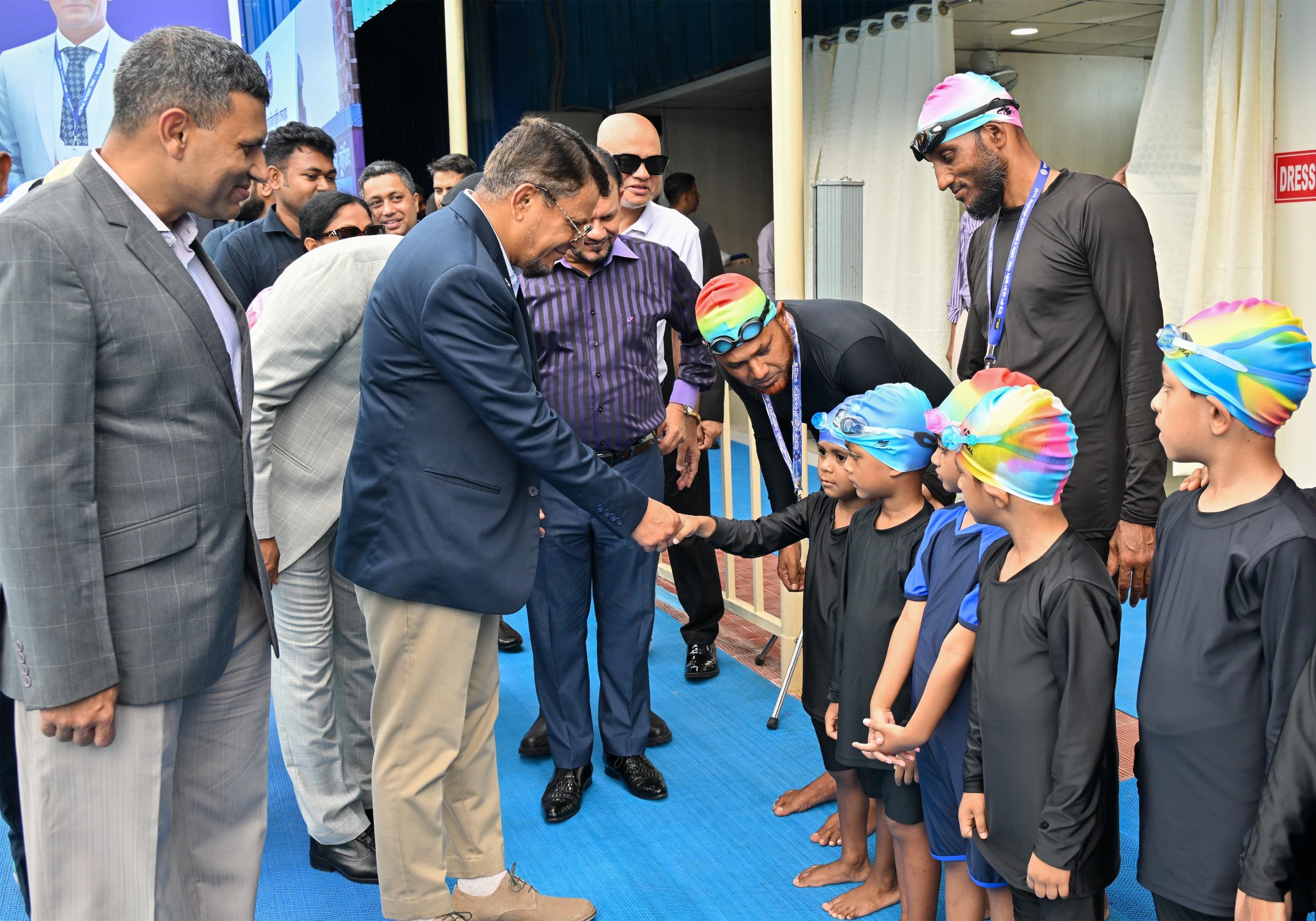 A guest of honor shakes hands with a young swimmer in a rainbow cap as coaches and children look on near the changing rooms.