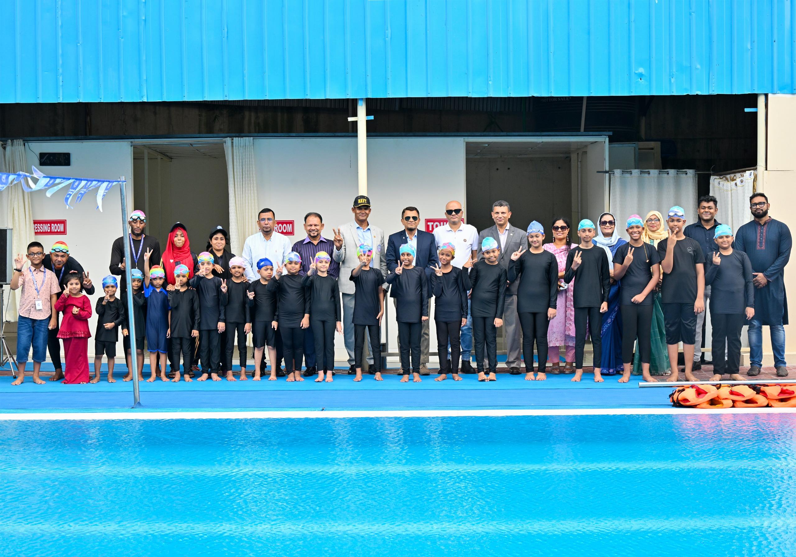 A large group of swimmers, coaches, and family members gather for a photo beside the changing rooms, a Bangladesh Navy cap visible among the guests.