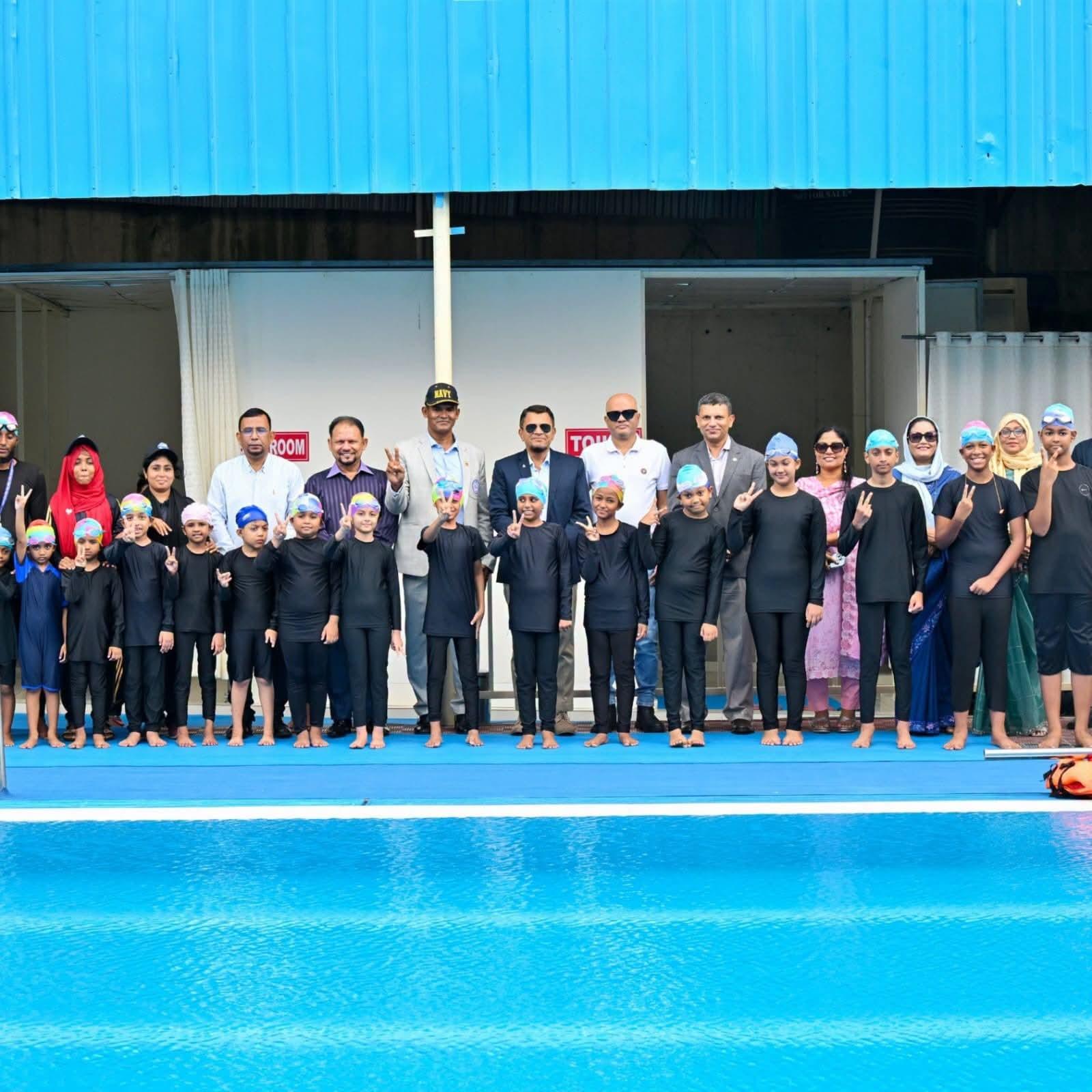 Coaches, officials, and young swimmers pose together on the pool deck, several flashing peace signs for the camera.