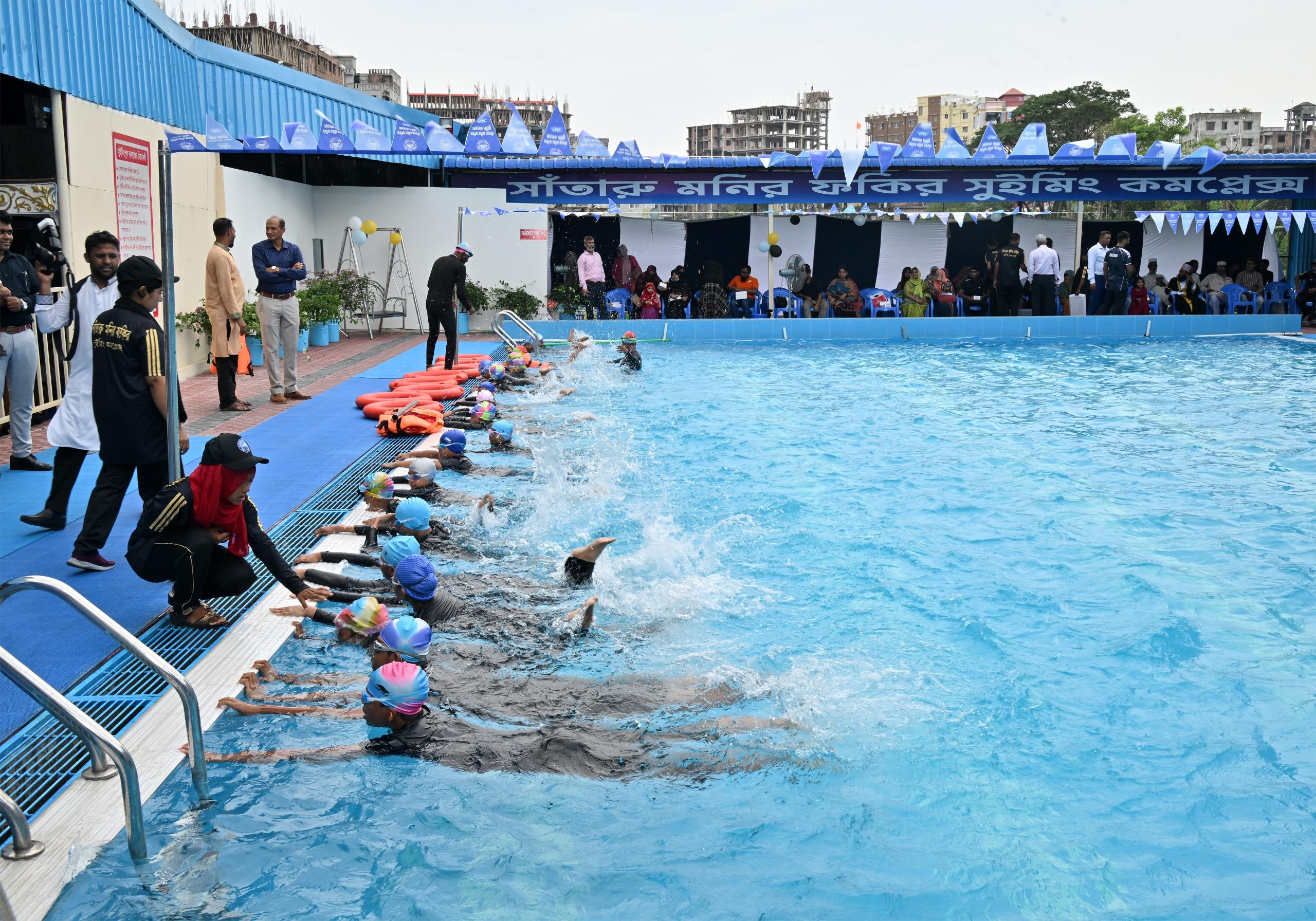 Beginner swimmers push off the wall together, spray catching the light, coaches crouched at the edge encouraging them.
