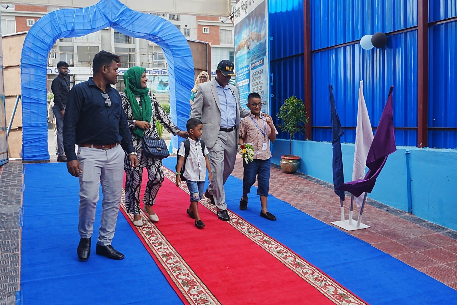 A family walks a red carpet through the blue balloon archway into the complex, a boy carrying a bouquet for the guest of honor.