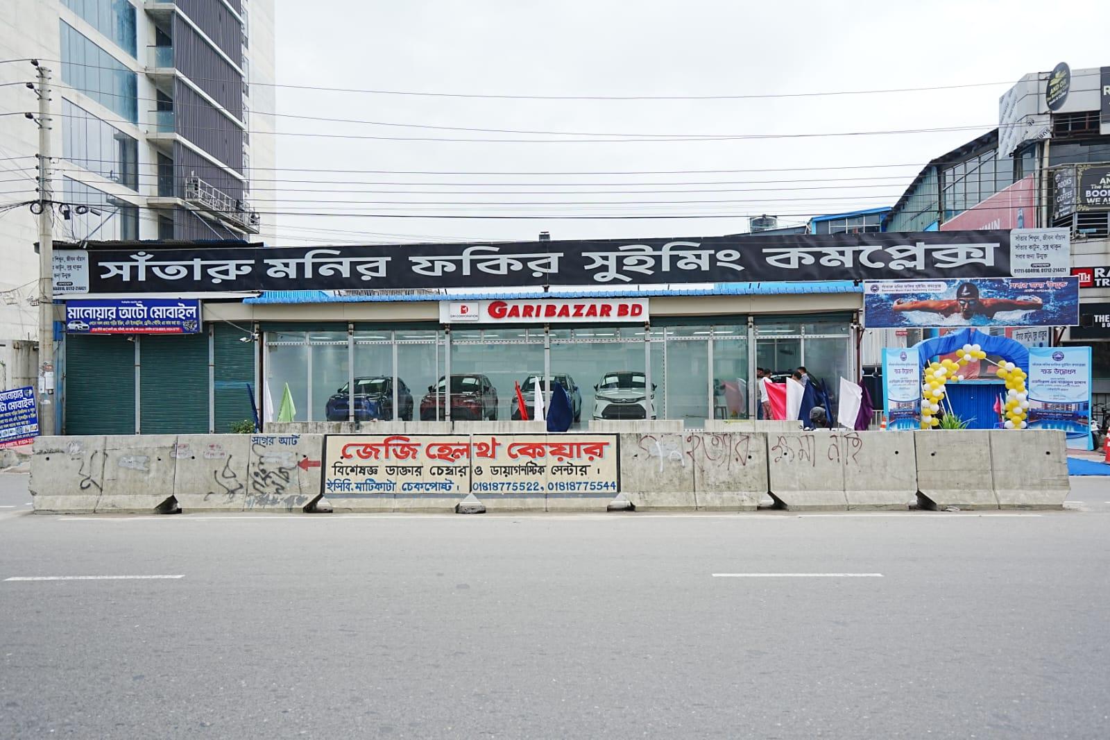 The street-level signage for Monir Fokir Swimming Complex in Bengali, mounted above a car showroom on ECB Chattar's main road.